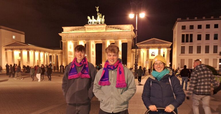 Drei junge Menschen stehen vor dem Brandenburger Tor und lächeln in die Kamera.