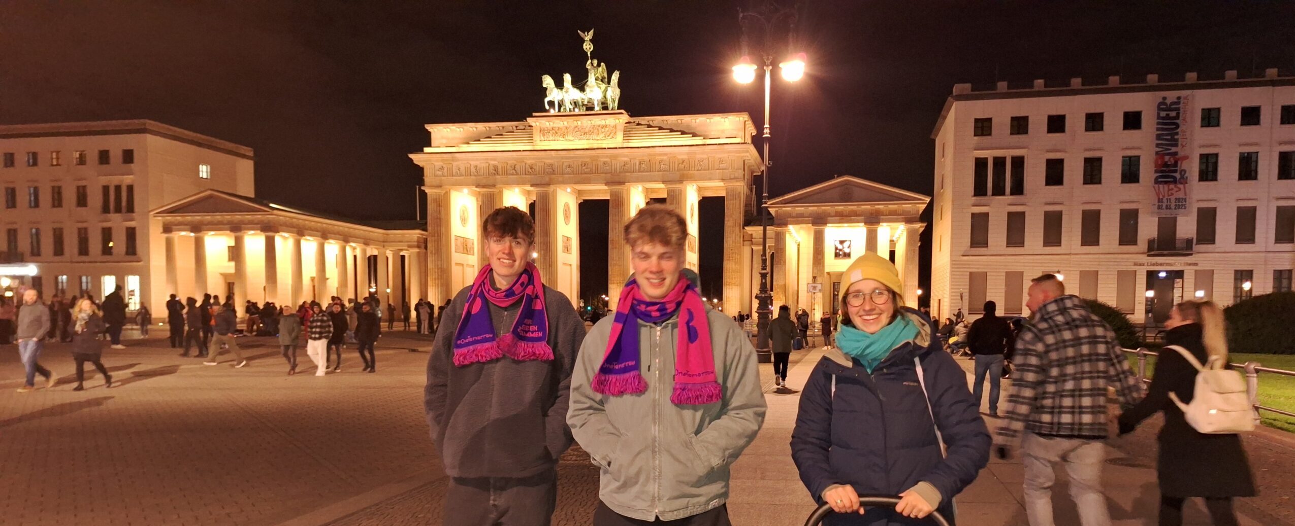 Drei junge Menschen stehen vor dem Brandenburger Tor und lächeln in die Kamera.