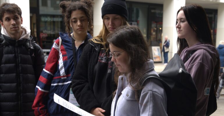 Eine junge Person steht in einer Fußgängerzone und liest ein Blatt Papier. Um sie herum stehen vier andere Junge Menschen.