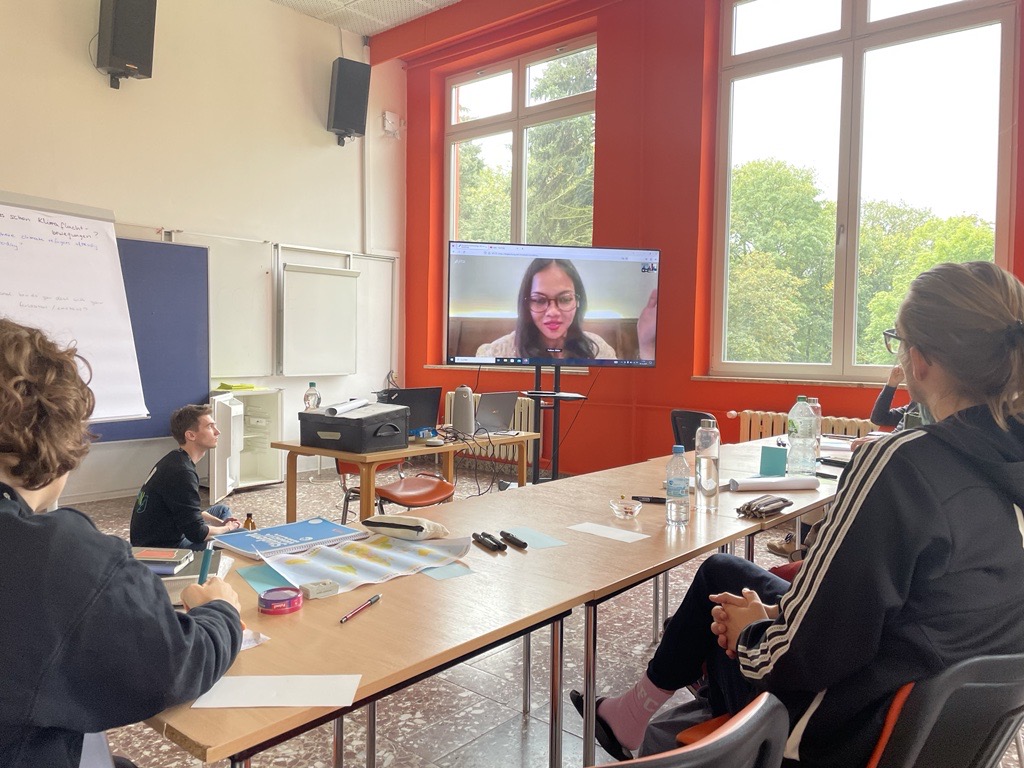 Mehrere junge Menschen sitzen in einem Seminarraum auf dem Boden oder vor Holztischen. Sie machen sich Notizen und schauen auf einen Bildschirm, auf dem eine junge Person mit Brille per Videokonferenz zu ihnen spricht.